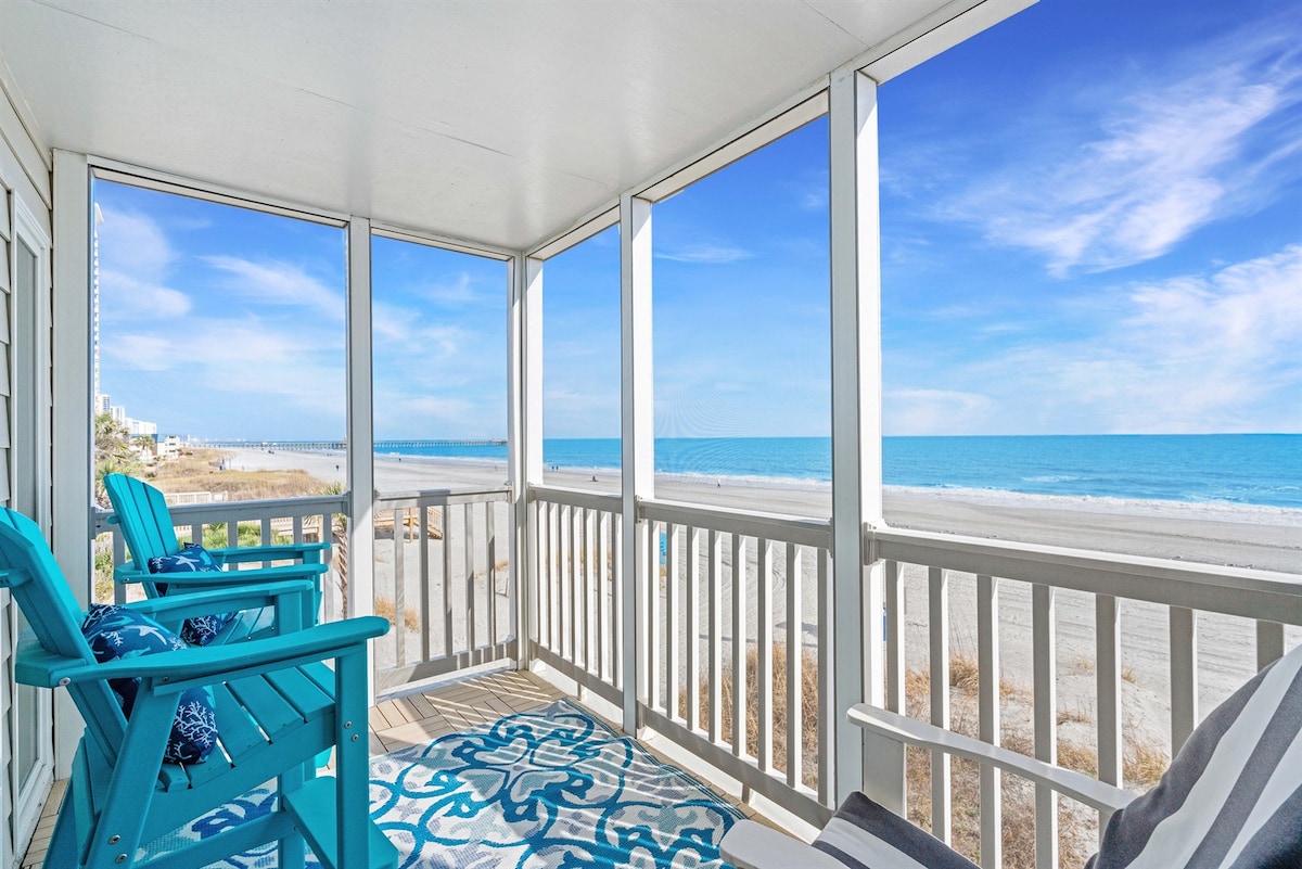 A screened-in balcony offers comfortable seating with two blue chairs, overlooking the sandy beach and ocean. The expansive view highlights the turquoise water under a bright sky, creating a serene coastal atmosphere.