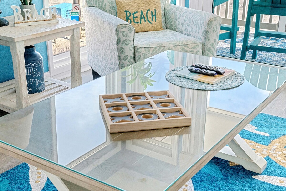 A stylish living area is highlighted by a glass coffee table featuring a wooden tic-tac-toe game. A light blue and white patterned chair displays the word 'BEACH.' Surrounding elements include a decorative blue vase and a round placemat on the table.