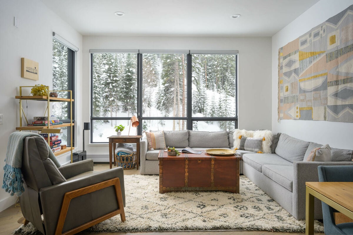 The living room features expansive windows that showcase snowy forest views. A large, soft rug covers the floor, while a sectional sofa and an armchair provide ample seating. A coffee table and a bright wall art piece complement the modern decor.