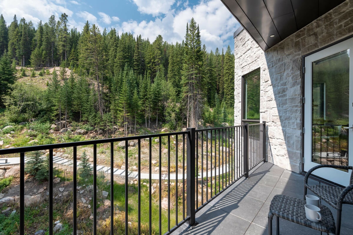A private deck is visible, featuring two cozy chairs and a small table. Beyond, a lush landscape of coniferous trees and rocky terrain creates a serene natural setting. Bright clouds float across a blue sky, enhancing the tranquil atmosphere.
