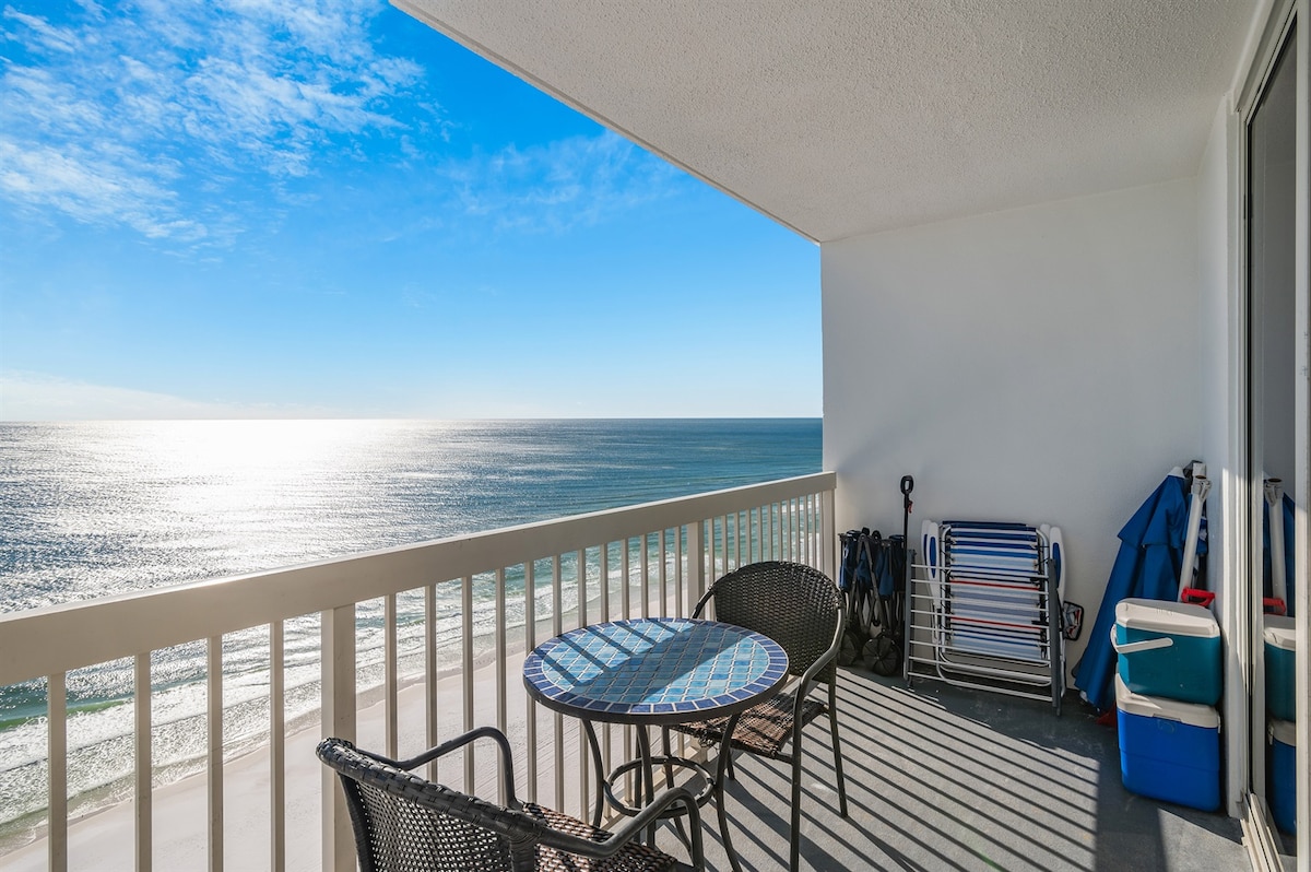 A balcony offers a stunning view of the Gulf, with the sun reflecting off the water. A small round table with two chairs is situated on the balcony, alongside a cooler and beach gear, enhancing the coastal atmosphere.