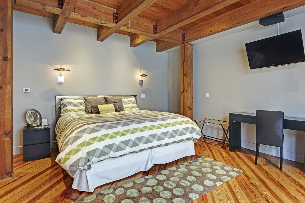 A spacious sleeping area is highlighted by a large bed with a patterned duvet cover. A flat-screen television is mounted on the wall, while a sleek desk and chair are placed nearby. Wood beams and polished flooring add warmth to the contemporary design.