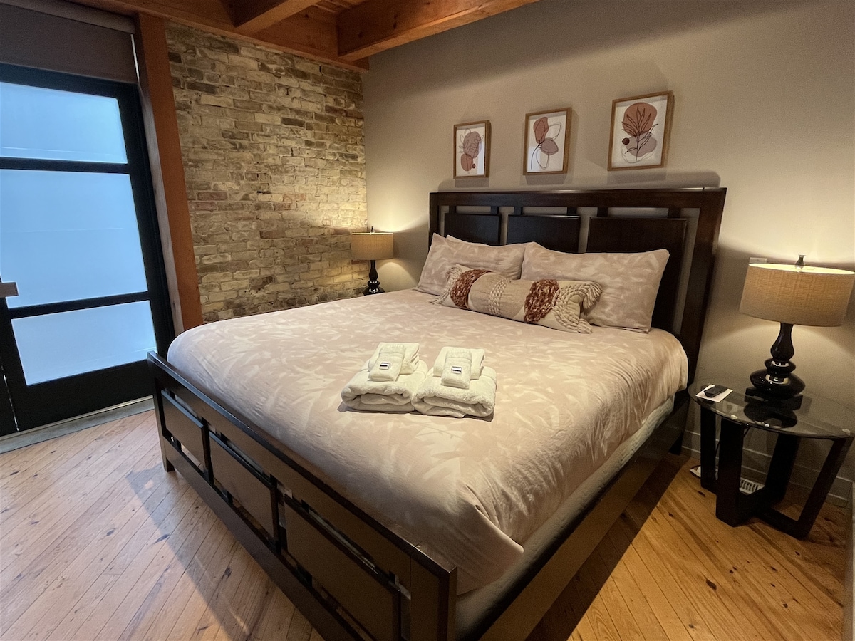 A spacious bedroom features a king size bed dressed in light-colored linens, flanked by bedside tables with lamps. A textured brick wall adds character, while three framed floral prints enhance the decor. Wooden beams highlight the ceiling, and sunlight enters through a nearby window.