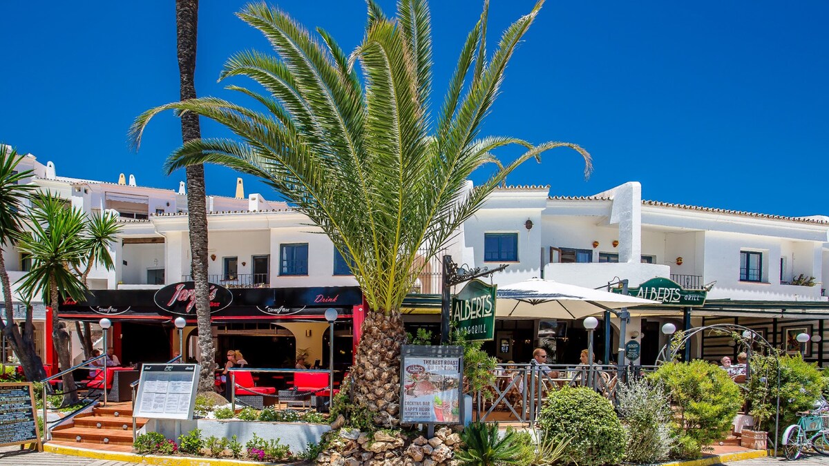 A vibrant scene showcases a palm tree in the foreground, framing a lively outdoor dining area. The buildings in the background reflect whitewashed architecture, complemented by bright blue skies. Tables are set under umbrellas, creating a welcoming atmosphere for patrons to enjoy their meals.