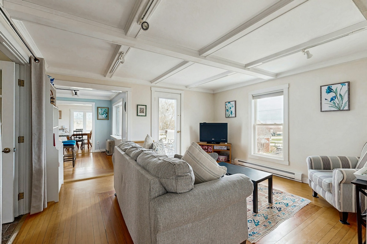 A spacious living area is framed by large windows, allowing natural light to fill the room. Comfortable seating includes a light-colored sofa and a striped armchair, with a coffee table centrally placed. Artwork adorns the walls, and a television is positioned for easy viewing.