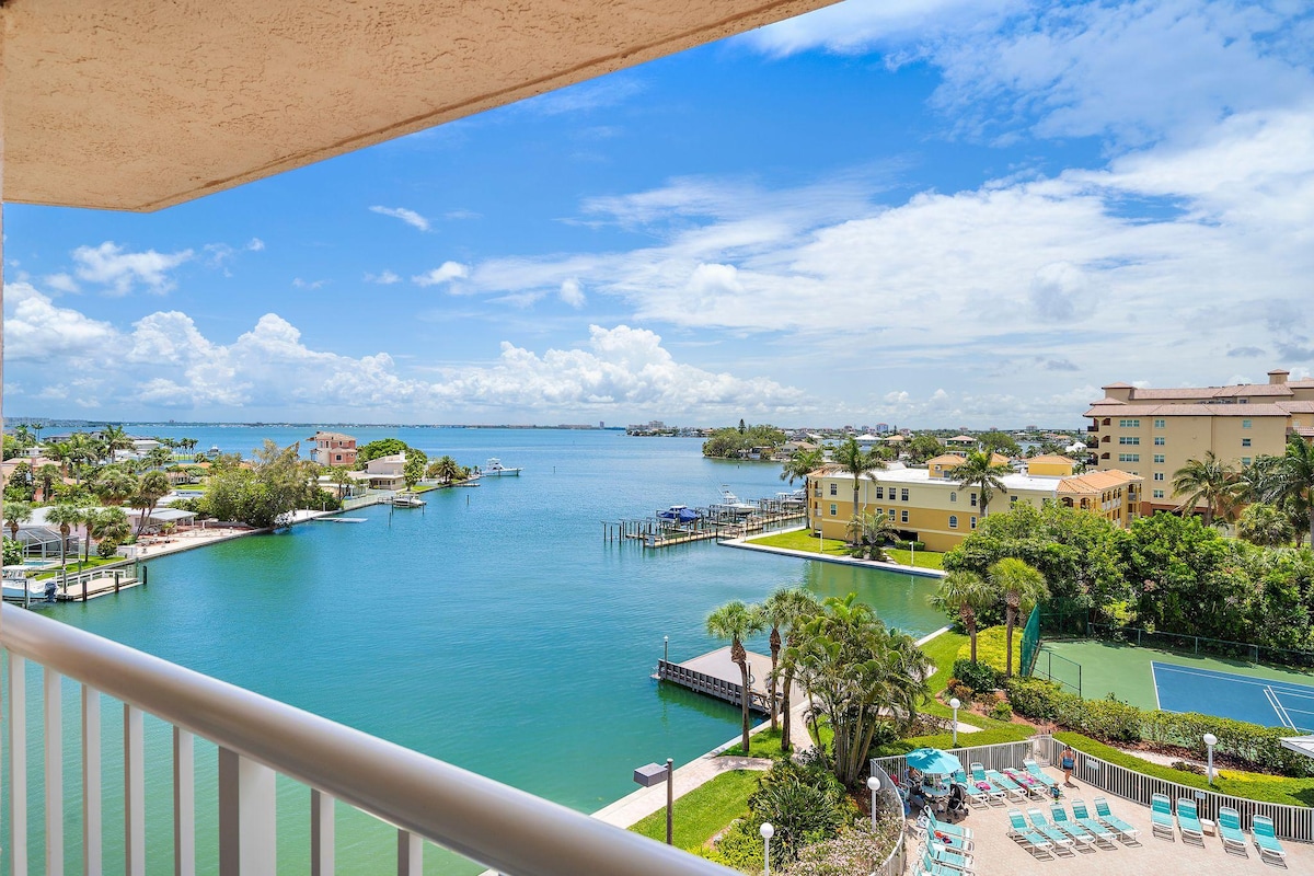 A scenic view from a balcony showcases the tranquil waters of Boca Ciega Bay, lined with lush greenery and picturesque homes. The image features a heated pool area and tennis courts, with clear blue skies enhancing the peaceful ambiance.