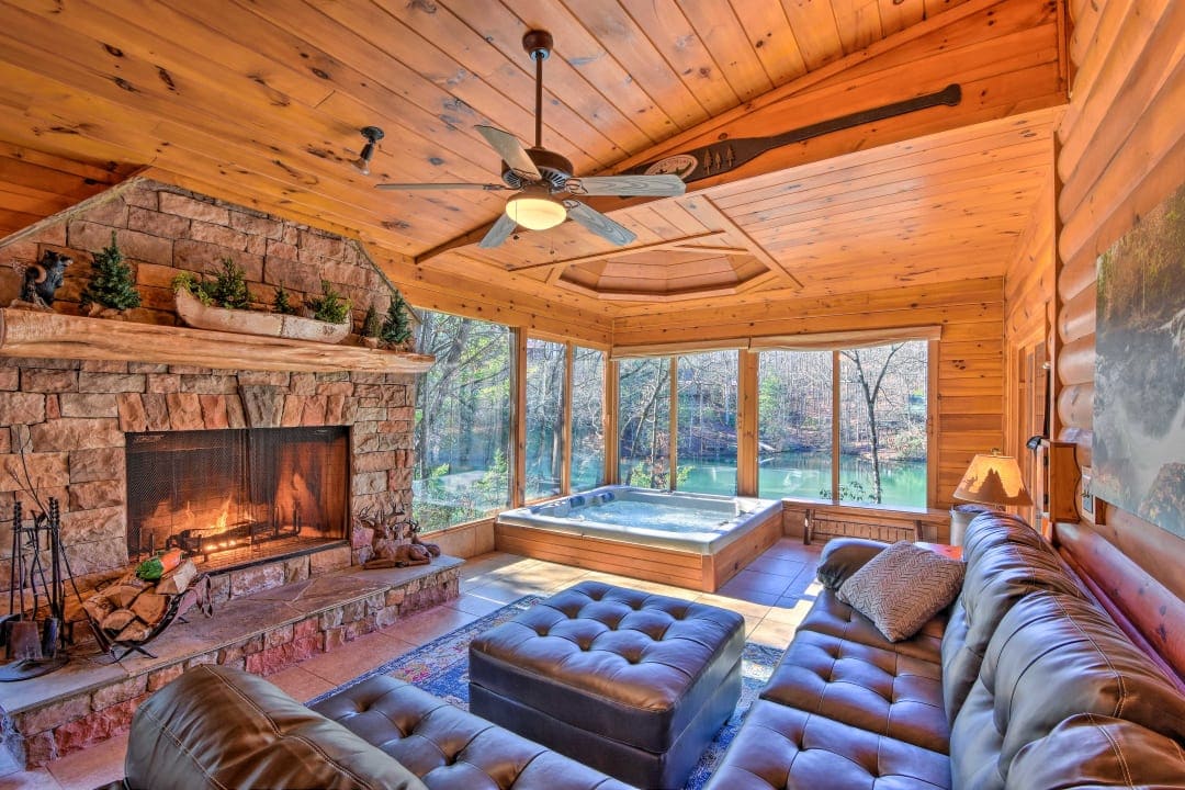 A cozy indoor living area features a spacious leather sectional sofa and a stone fireplace. Large windows provide views of the surrounding nature, while a hot tub is positioned nearby, creating a relaxing atmosphere. The wooden ceiling accentuates the cabin's charming design.