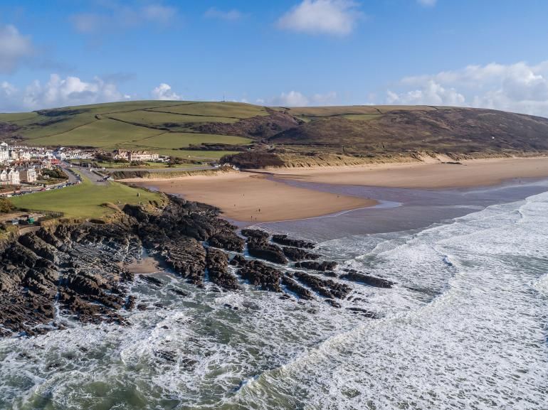 A sweeping view captures the expansive sandy beach bordered by rocky formations, with gentle waves lapping at the shore. Hills covered in greenery rise in the background under a clear blue sky, creating a serene coastal landscape.