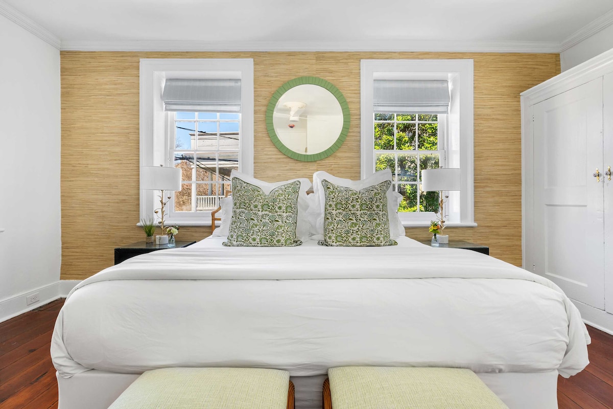 A bright bedroom features a spacious bed adorned with decorative pillows. Light filters through two windows, highlighting the natural textures of the walls. A round mirror adds a touch of elegance, while a pair of potted plants sit on the bedside tables.