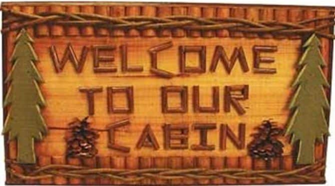 A wooden sign with the words 'Welcome to Our Cabin' is prominently displayed. The sign features carved decorative elements, including two stylized pine trees and pinecones, framed by a rustic wooden border.