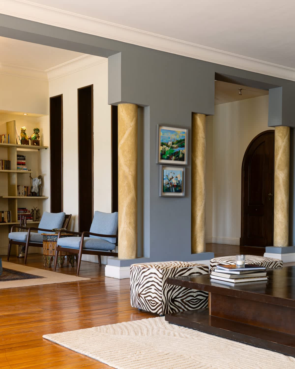 A stylish living space features elegant columns with gold accents and a section for seating with two blue chairs. A small coffee table is centered on a patterned area rug, complemented by a wooden console with decorative items and two framed artworks on the wall.
