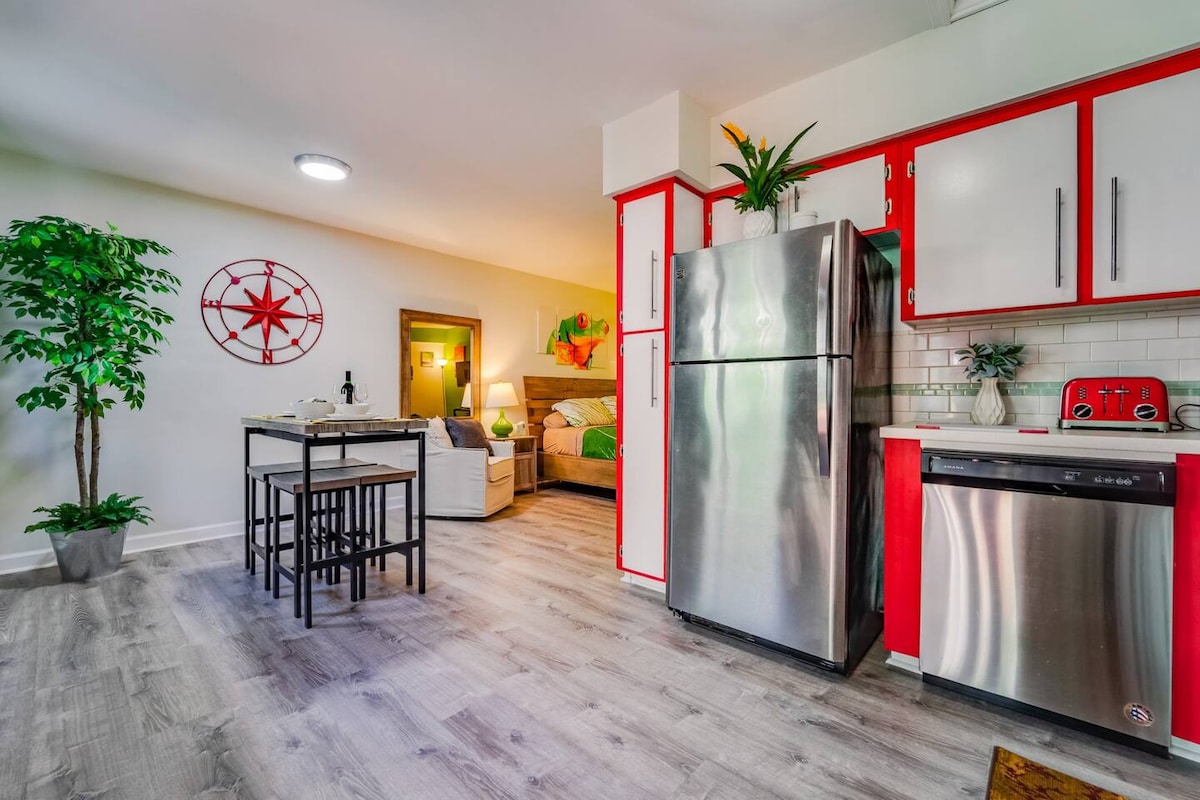 The kitchen area displays red and white cabinets, complemented by stainless steel appliances. A dining table with seating is positioned in the open space. A frosted glass door leads to a cozy bedroom in the background, which is illuminated by soft lighting.