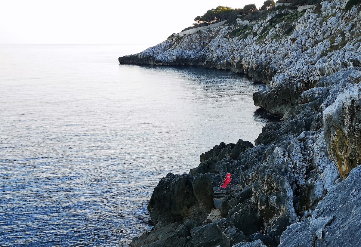 A rocky coastline extends along the clear, calm waters of the Lower Adriatic Sea. The smooth, grey rocks contrast with a vibrant red sun lounger placed on a flat surface. The distant shoreline features hills covered in greenery, providing a serene backdrop.