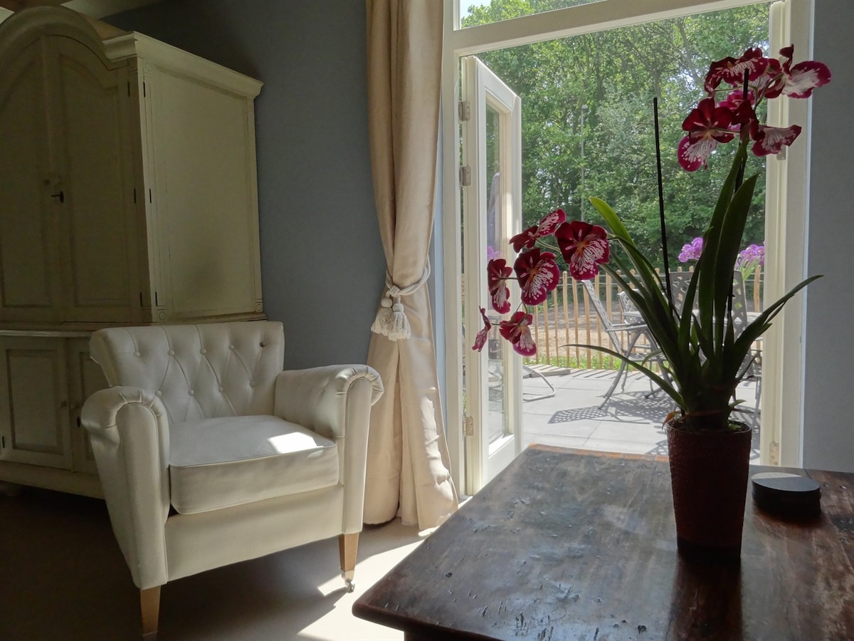 A comfortable white armchair is positioned beside a wooden table, adorned with a potted orchid. Large glass doors open to a view of the outdoor area, allowing natural light to brighten the room. Soft, neutral tones are present in the decor.
