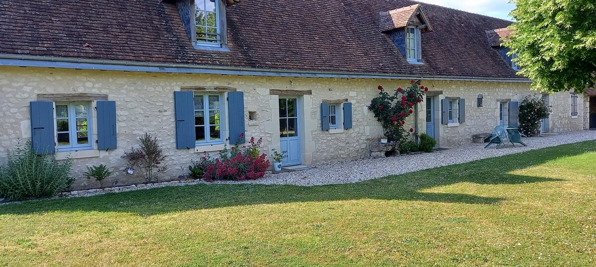 The exterior of the charming longère is showcased, featuring light-colored stone walls and traditional blue shutters. A well-maintained garden is visible, adorned with vibrant flowers and greenery, while a gravel pathway leads up to the main entrance, inviting visitors into the home.