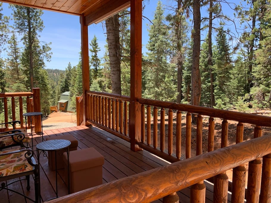 A covered porch features natural wood beams and railings, providing a relaxing space with rustic chairs and a small table. The view showcases surrounding dense pine trees under a clear blue sky, inviting guests to enjoy the peaceful outdoor setting.
