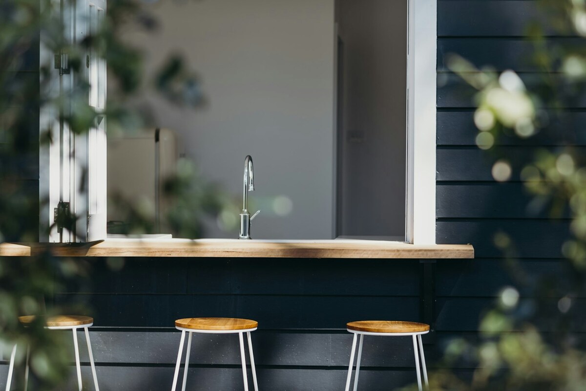 A window with a wooden countertop is framed by greenery, revealing a clean, modern kitchen interior. Three wooden stools are positioned neatly beneath the counter, contributing to an inviting and functional outdoor space.