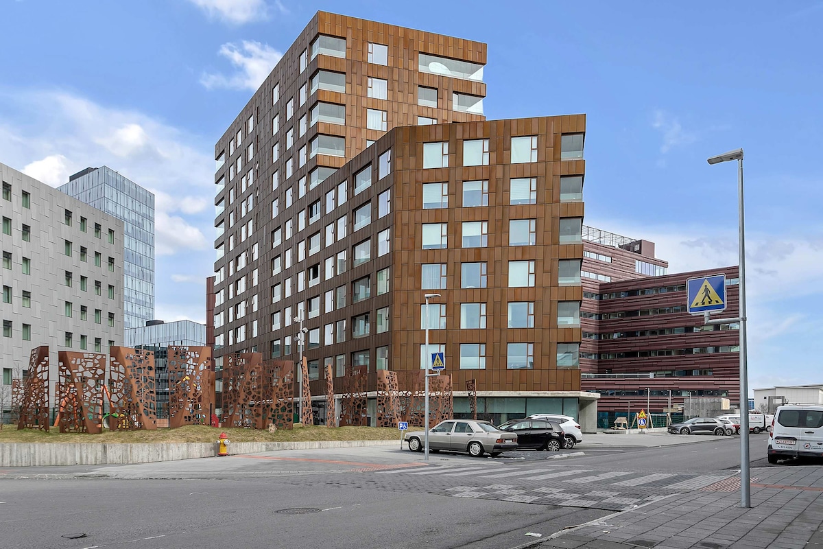 The modern Sif Apartments complex features a distinctive architectural design with a combination of metal and glass facades. The building is situated in an urban environment, with surrounding greenery and parking visible in the foreground, creating a contemporary and inviting atmosphere.