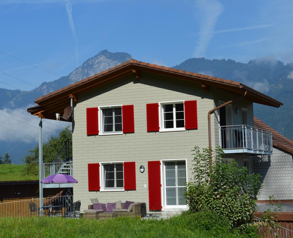 The farmhouse with a light grey exterior features red shutters and a covered balcony. Outdoor seating is provided under an umbrella, with a sofa set positioned in front of the house. The surrounding landscape includes rolling hills and a mountainous backdrop.
