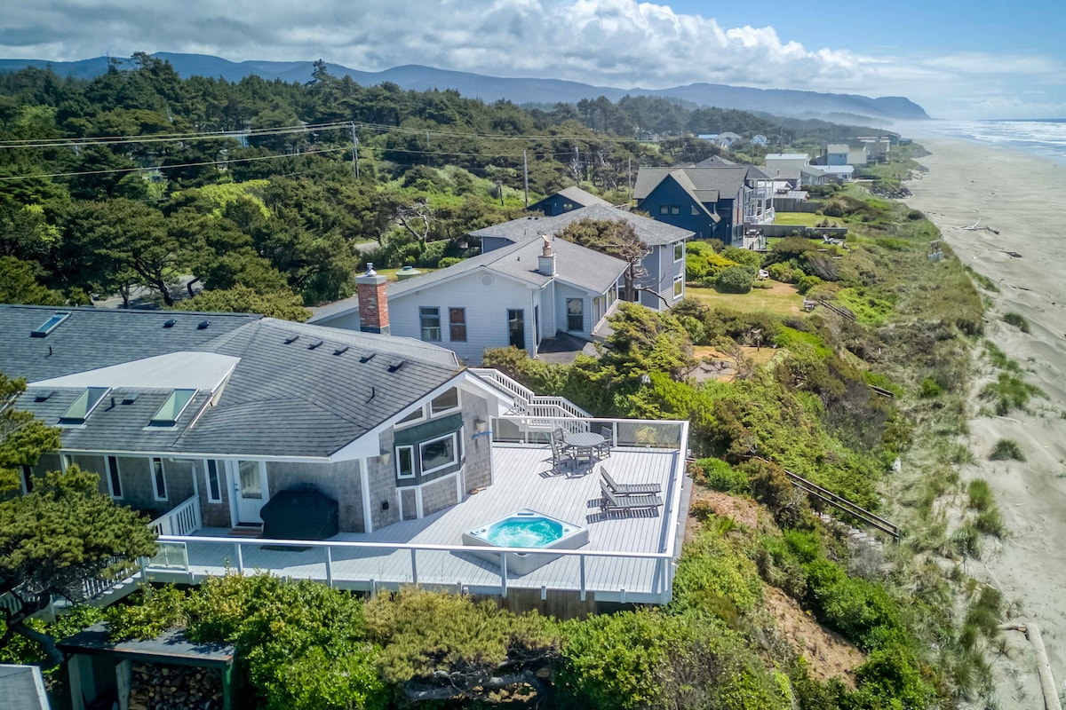 An aerial view captures a stylish oceanfront home with multiple decks overlooking the Pacific Ocean. The hot tub is visible on the deck, complemented by outdoor seating. Lush greenery surrounds the property, with sandy beach and coastal hills in the background.