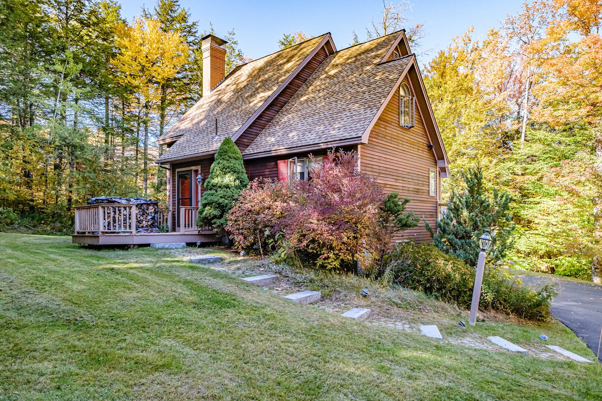 The exterior of the home features a charming brown shingle facade with a steeply pitched roof. Surrounding lush greenery is complemented by vibrant fall foliage. A spacious deck with a patio area is visible, along with a well-kept lawn leading to a stone pathway.