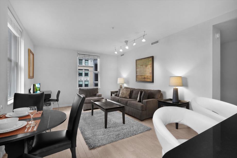 A spacious living area includes a sectional sofa and matching armchairs. A coffee table centers the room, while a dining table is set for four. Two lamps provide soft lighting, and large windows allow natural light to fill the space.