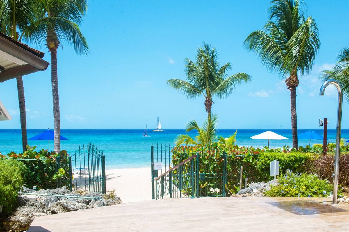The image depicts a vibrant beach scene with clear turquoise waters and a sailboat in the distance. Lush palm trees frame the view, and umbrellas provide shade along the shoreline. A private gate leads to the beach, enhancing the tranquil atmosphere.