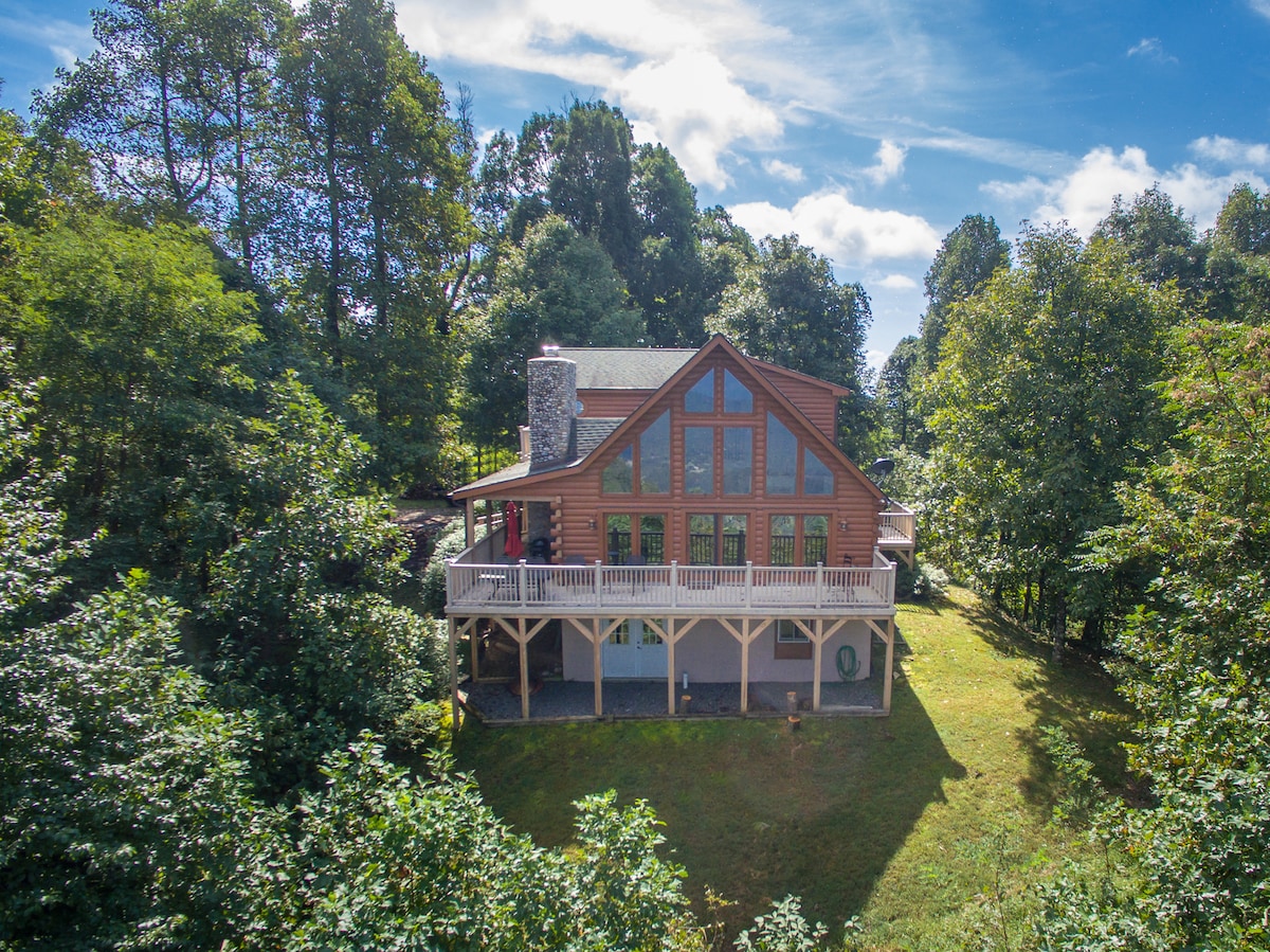 A spacious cabin is nestled among trees, showcasing a wooden exterior with large windows. The elevated porch offers views of the surrounding landscape. The roof features a gabled design, complemented by stone accents at the corner, creating a welcoming outdoor space.