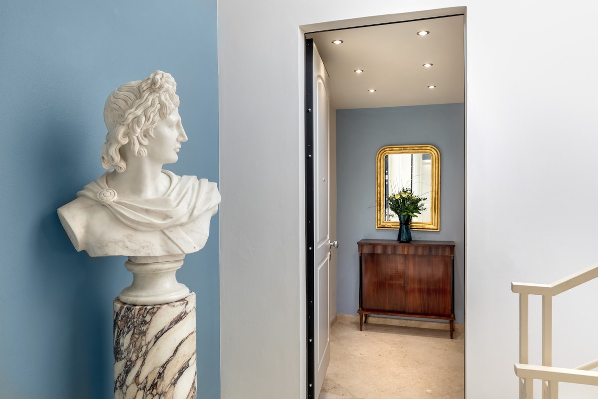 A hallway is displayed, featuring a classical marble bust positioned on a column. The walls are painted in a soft blue tone. A decorative chest stands against the opposite wall, complemented by a mirror above it. A floral arrangement sits in a vase on the chest.