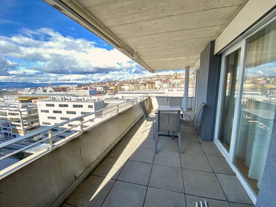 A spacious balcony offers a panoramic view of the city skyline, set against a backdrop of partly cloudy skies. A small table and two chairs are positioned on the tiled surface, providing an inviting space to relax outdoors.