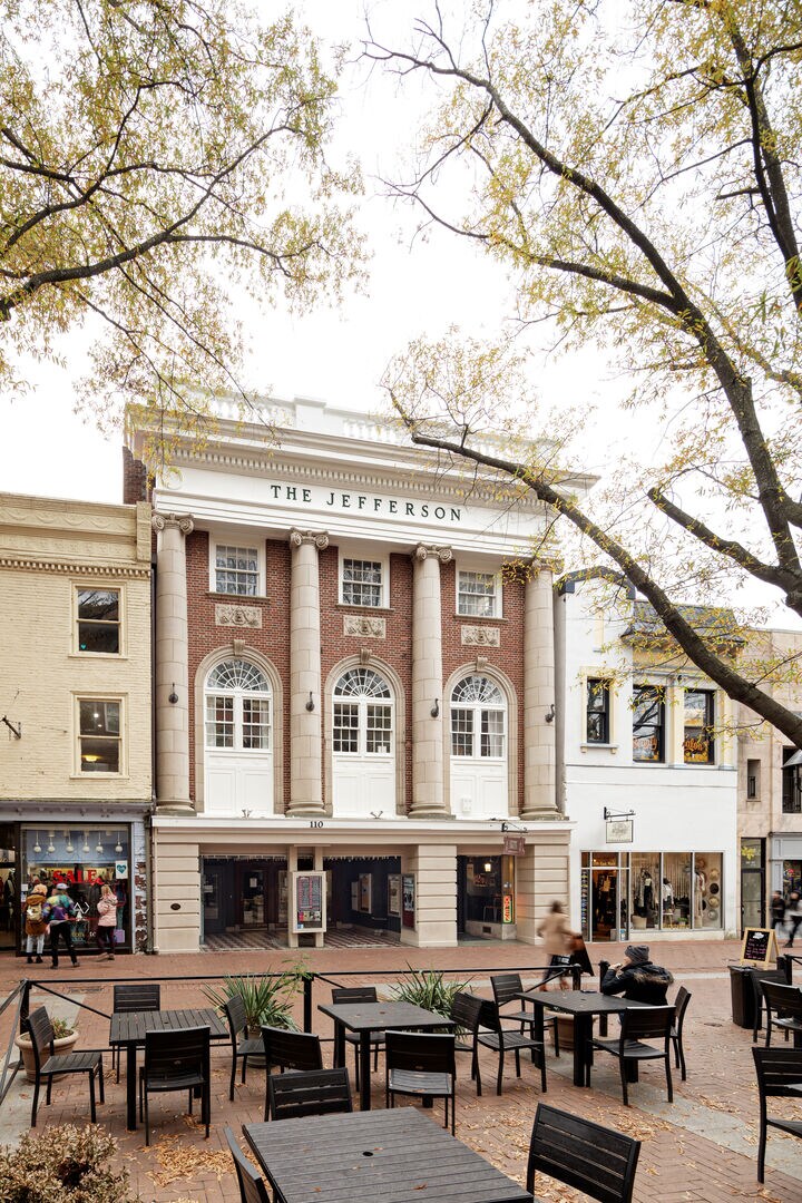 The historic facade of The Jefferson is showcased, featuring classic architectural elements such as tall columns and large windows. Outdoor seating is arranged on a pedestrian mall, surrounded by trees, with shops visible in the background, enhancing the vibrant atmosphere.