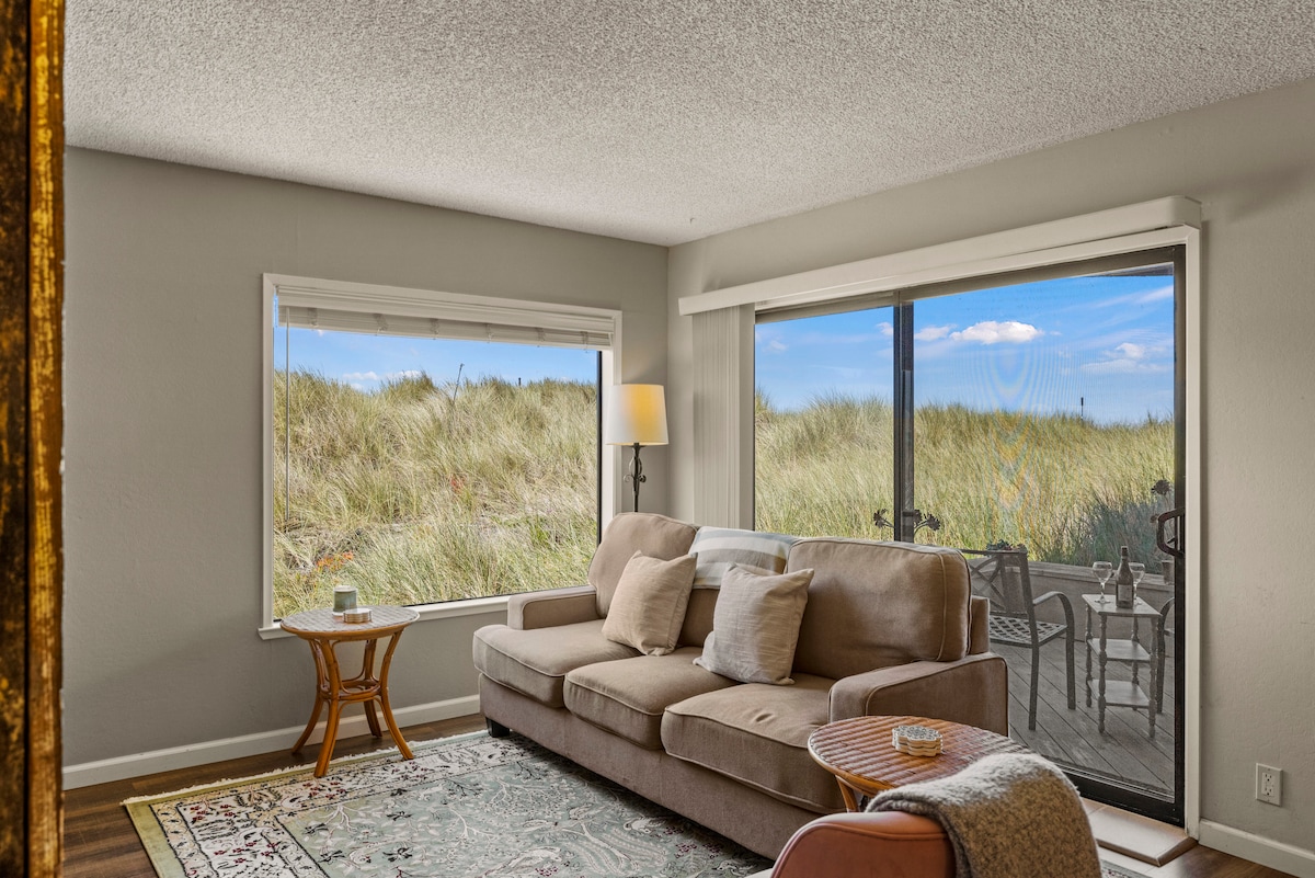 A living area is presented with natural light streaming through large windows, framed by tall dune grass. A comfortable sofa faces the view, accompanied by a side table and a lamp, while a cozy rug adds warmth to the space.