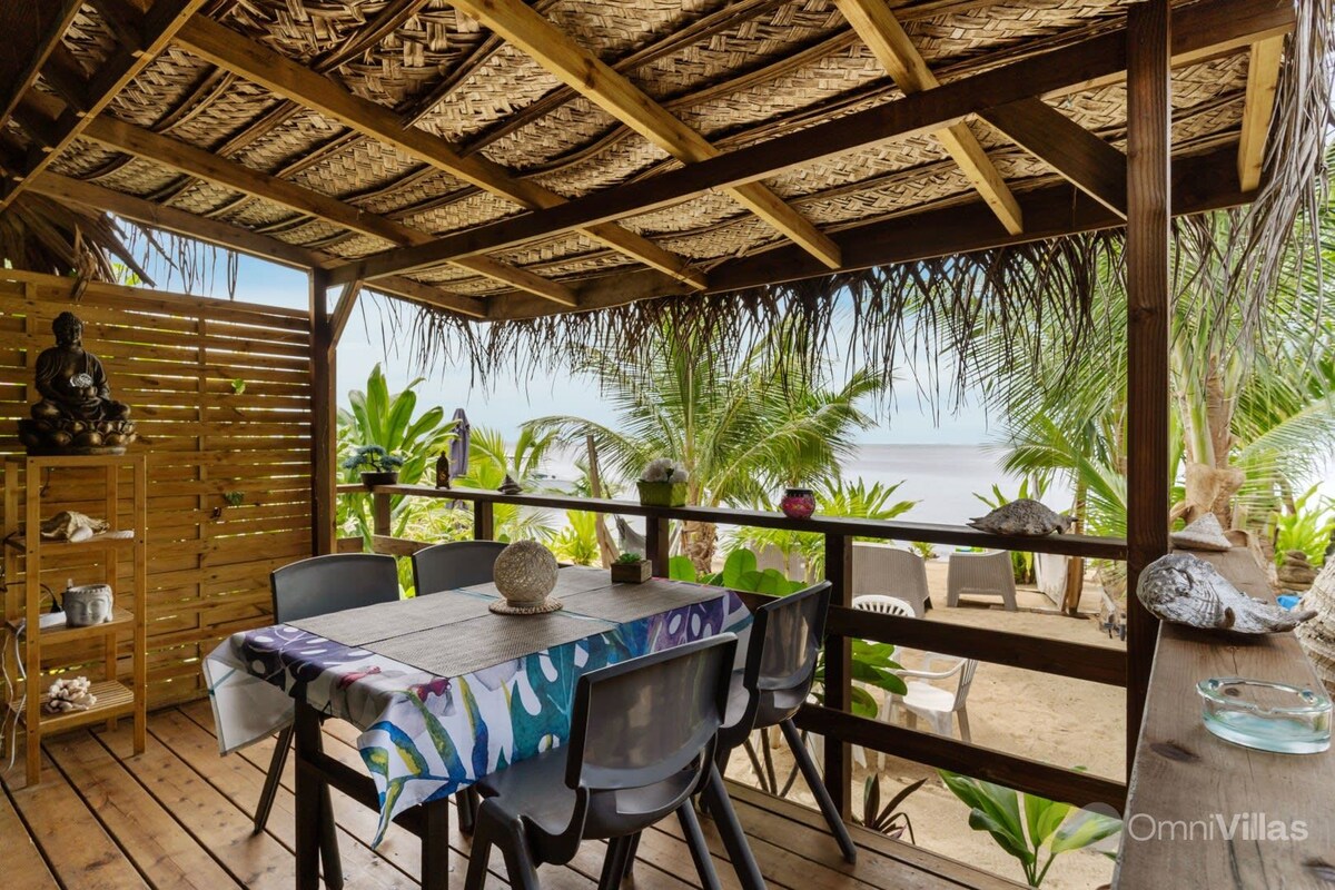 A shaded terrace features a wooden dining table surrounded by chairs. Lush tropical plants frame the view of the seaside. Natural materials, such as woven ceiling panels, complement the relaxed outdoor setting, providing a harmonious connection with the surrounding landscape.