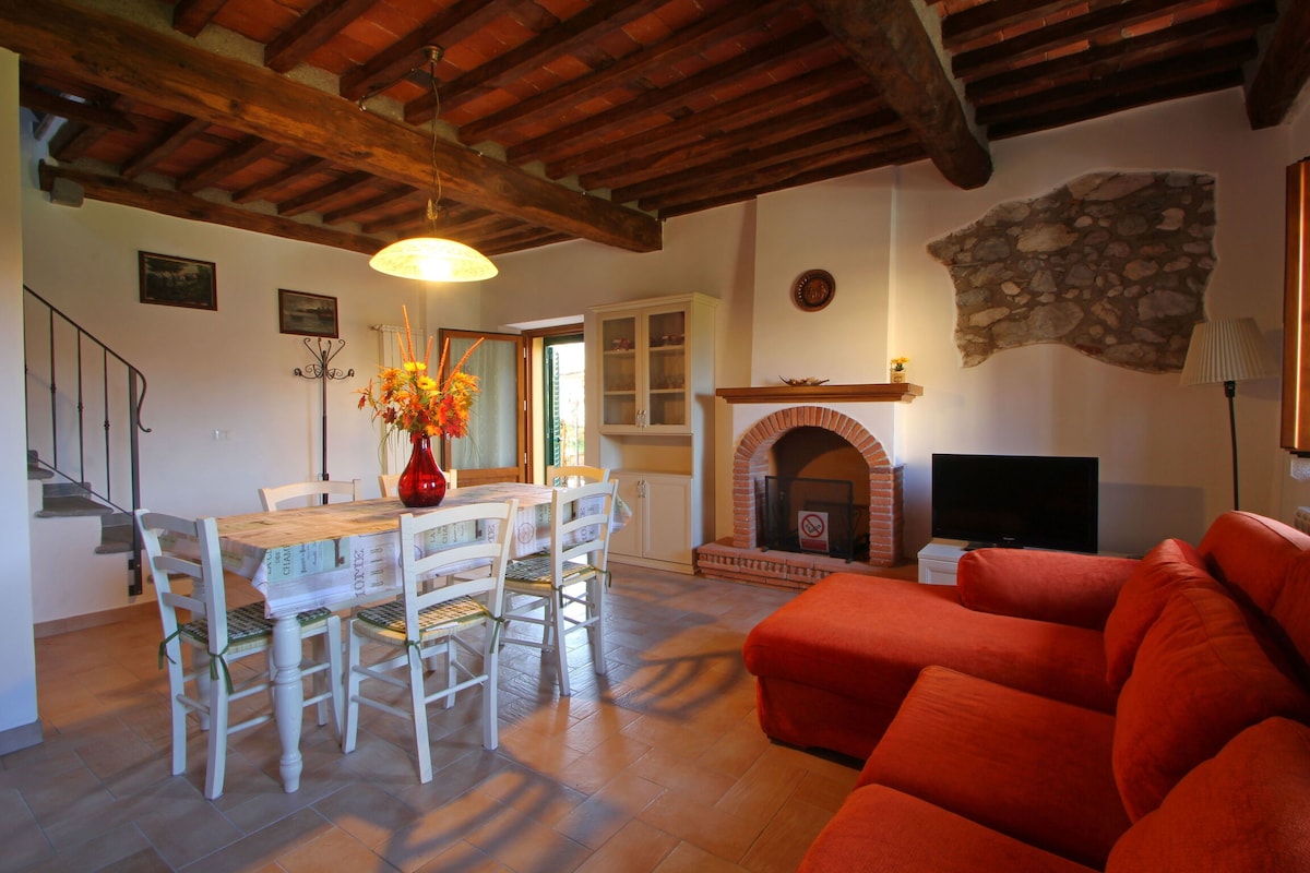 A living room features a comfortable red sofa, a wooden dining table with six chairs, and a TV beside a decorative fireplace. The space is illuminated by a pendant light, and a doorway leads outside, enhancing the cozy ambiance of the rustic interior.