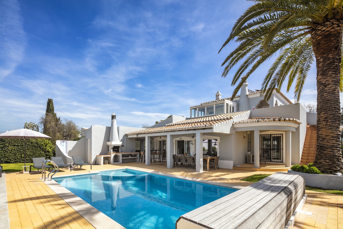 The villa features a large outdoor area with a private heated saltwater pool surrounded by sun loungers and a shaded dining space. Lush palm trees add to the tropical ambiance, while the outdoor barbecue area is visible alongside the spacious terrace.