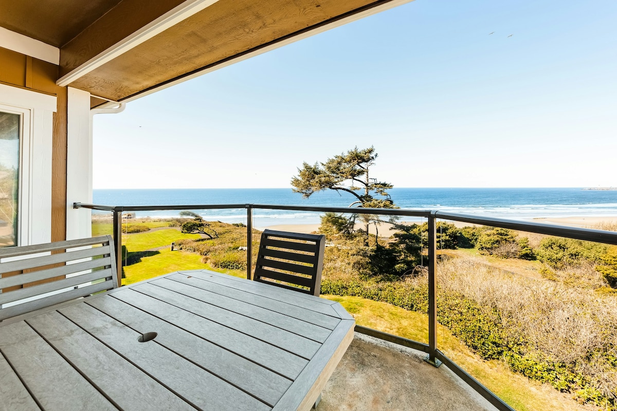 A balcony offers a table and chairs with a view overlooking the ocean and sandy beach. The scenic landscape features greenery and a solitary tree. Clear blue skies stretch above, complementing the tranquil ocean waters in the distance.