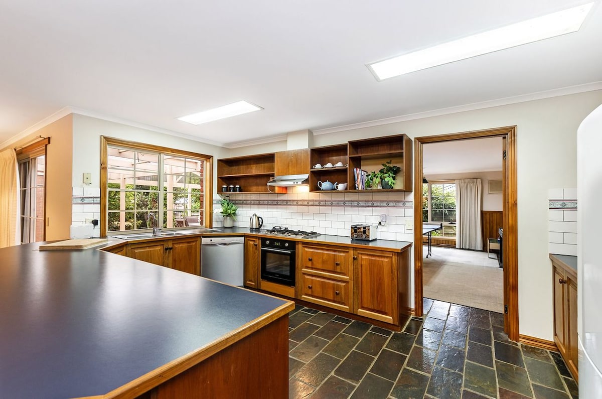 The kitchen features a spacious layout with wooden cabinetry and a dark countertop. Appliances include a stove and dishwasher. Natural light enters through large windows, illuminating the area. Adjacent doorways lead to the dining and living spaces, enhancing convenience for meal preparation.