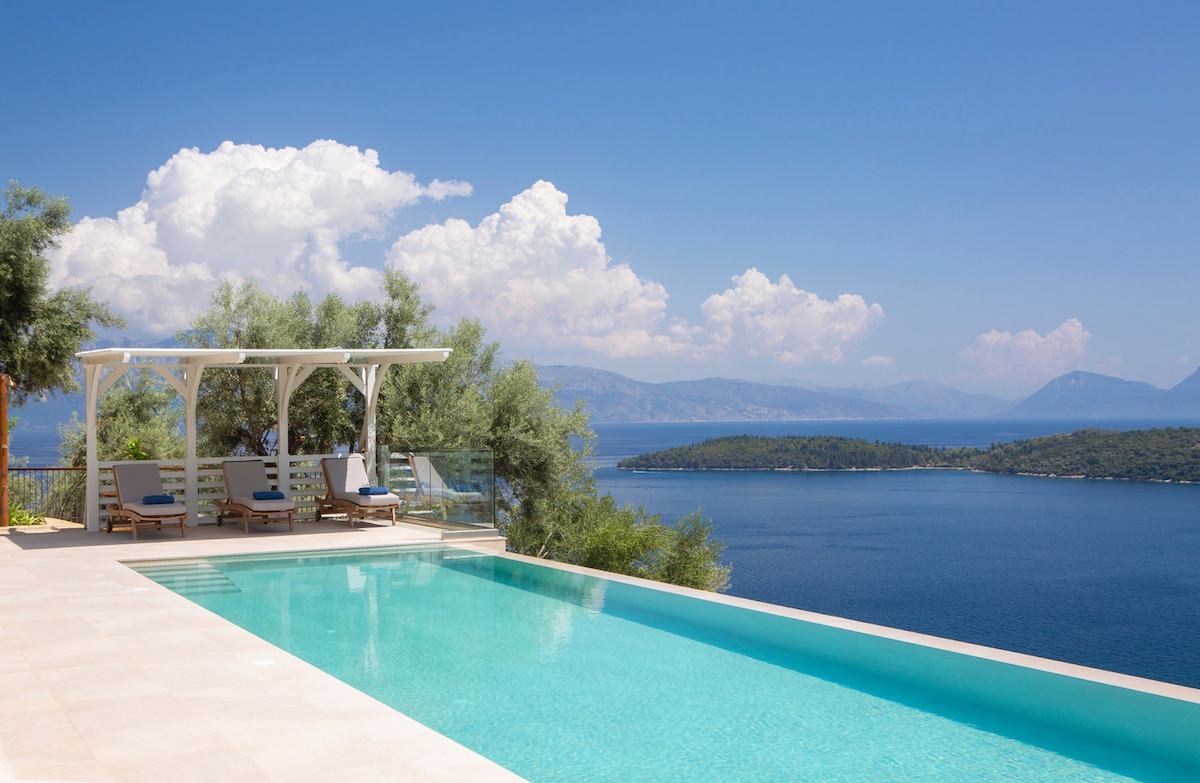 A long, heated swimming pool stretches across the patio, surrounded by olive trees. A shaded terrace features lounge chairs, offering sweeping views of the sea and distant islands under a bright blue sky with scattered clouds.