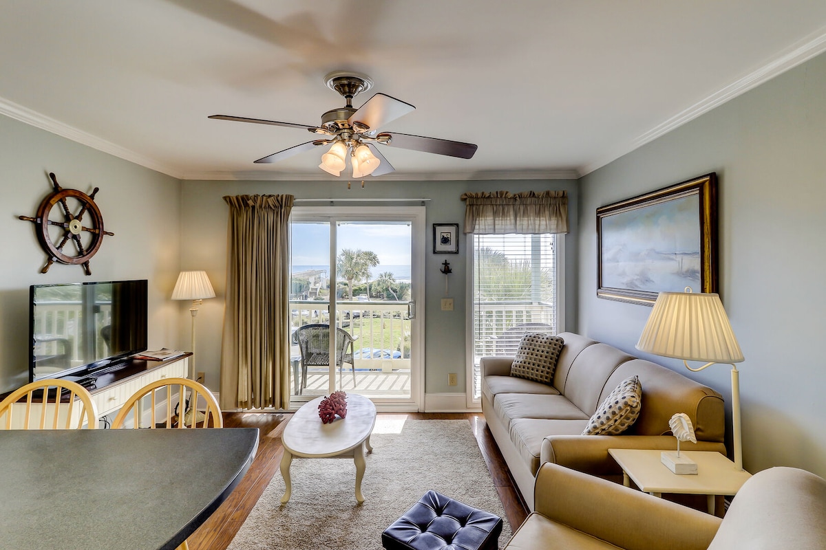 A well-lit living area includes a comfortable sofa and a TV stand, with large windows offering a view of the ocean and outdoor space. Decorative nautical accents contribute to the coastal theme, while a ceiling fan provides additional comfort.