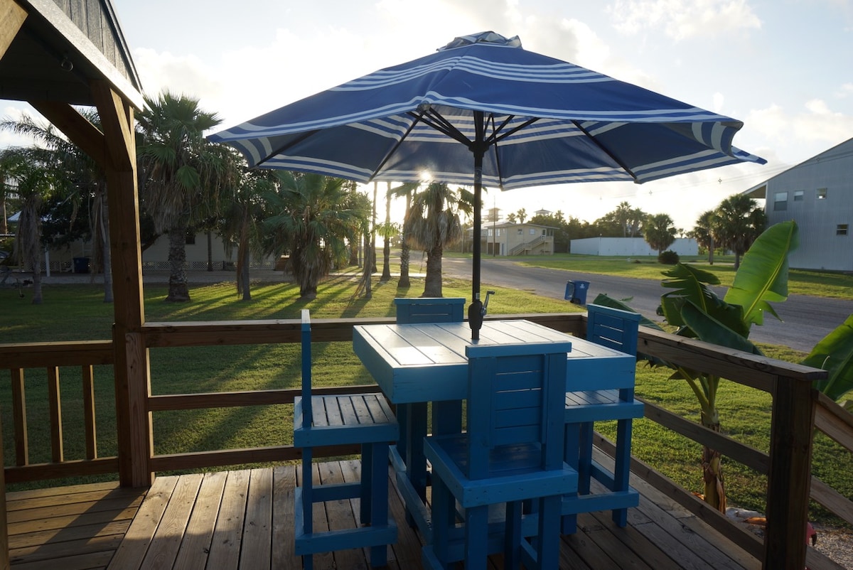 An oversized deck features a vibrant blue table set under a large umbrella, providing shade. Surrounding palm trees and greenery create a natural setting, while a gentle sunset casts a warm light across the outdoor space.