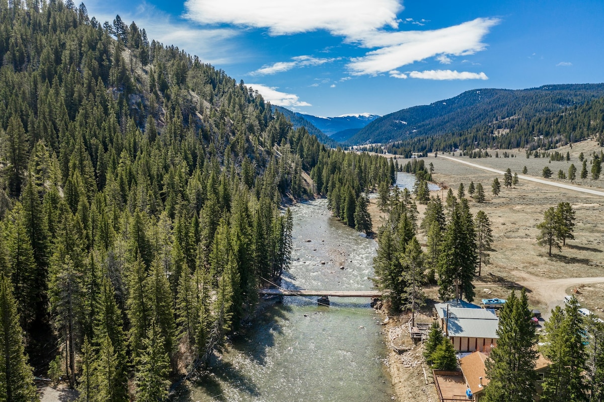 An aerial view captures the Gallatin River flowing through a scenic landscape surrounded by dense evergreen trees and rolling hills. The tranquil waters reflect the sky, with mountains visible in the distance under a partly cloudy sky.