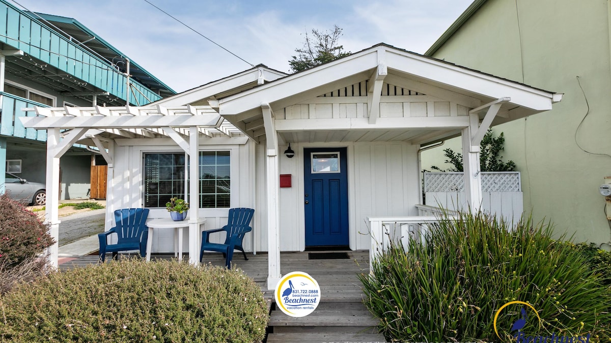 The exterior of a charming beach cottage is depicted, showcasing a white façade with a pitched roof and blue front door. A small table and two blue chairs are positioned on a wooden deck, surrounded by lush greenery and low shrubbery.