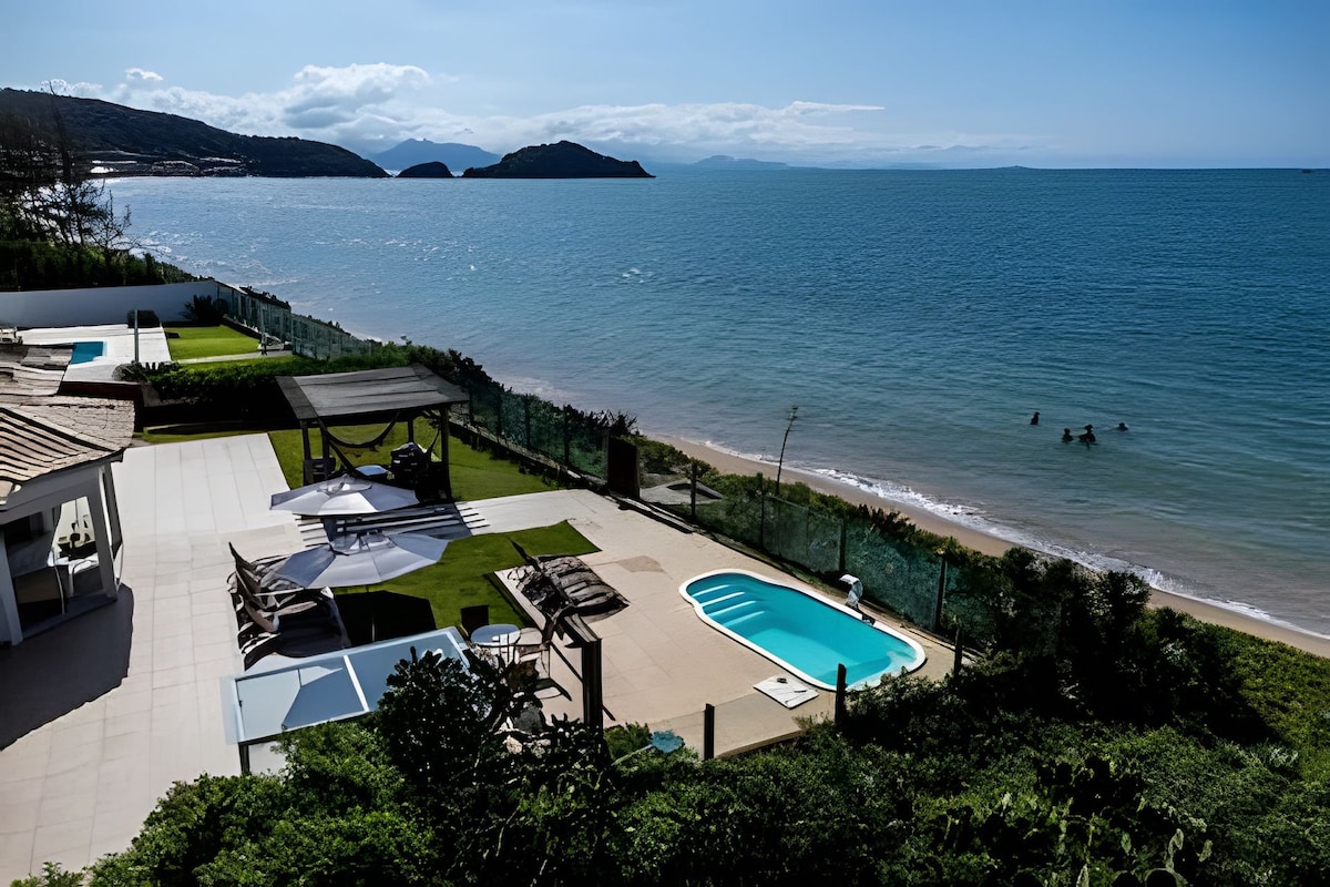 A serene beachfront view displays a private pool beside a sunlit patio. Loungers and a shaded area invite relaxation, while gentle waves lap the shore nearby. The expansive ocean stretches toward distant islands under a clear blue sky.