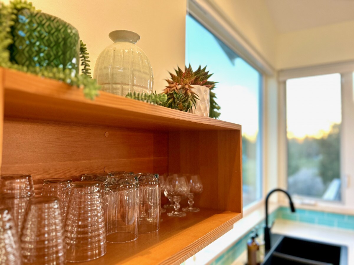 A wooden shelf holds an assortment of glassware, including tumblers and stemmed glasses, arranged neatly. Decorative plants and a small glass vessel are placed above the glassware, while a large window in the background provides a view of the outdoors.