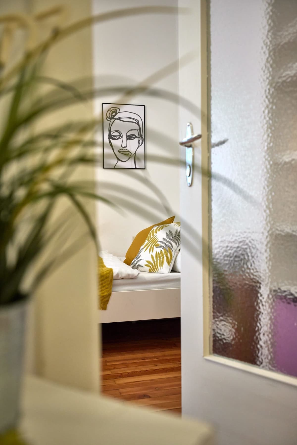 A view through a half-open door reveals a cozy bedroom space featuring a white bed adorned with decorative pillows in yellow and green tones. Soft wooden flooring complements the tranquil ambiance, while a stylish wall art piece adds a touch of elegance.