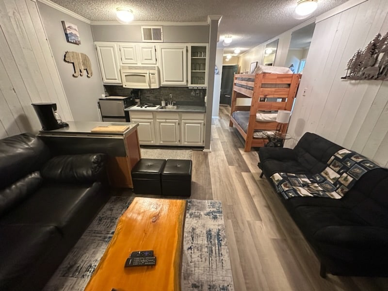 A living area features a black leather sofa alongside a futon, with a wooden coffee table positioned centrally. A kitchenette is visible, equipped with a stove and refrigerator. Built-in bunk beds are seen in the rear, providing sleeping space for children.
