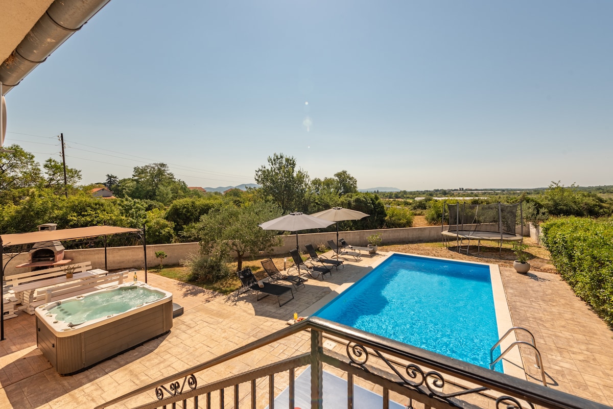An outdoor area showcases a large swimming pool and a hot tub, surrounded by lounge chairs and shaded by umbrellas. Lush greenery and trees provide a natural backdrop, enhancing the space's privacy. A clear blue sky and distant hills are visible in the background.