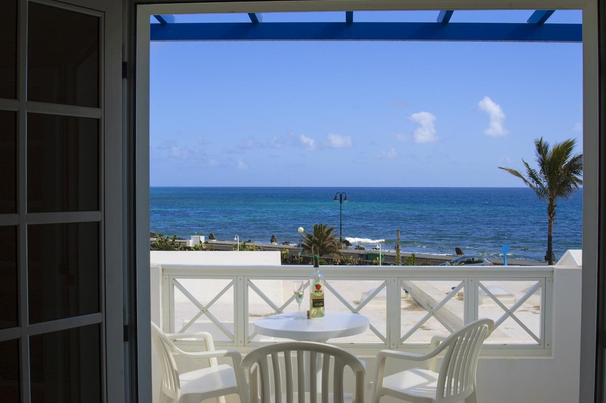 A view of the ocean is framed by an open balcony door. White outdoor furniture, including a round table and two chairs, is positioned on the balcony. Clear blue skies and gentle waves create a serene coastal atmosphere.