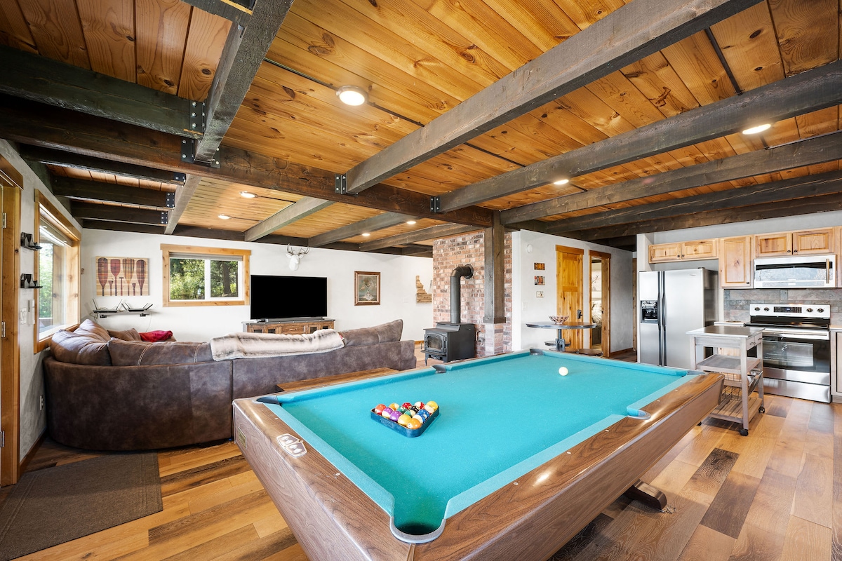 An inviting game room features a teal pool table positioned centrally. Natural light is provided by windows, illuminating the rustic wooden beams and walls. A comfortable couch and a flat-screen TV are visible, creating a casual space for relaxation and entertainment.