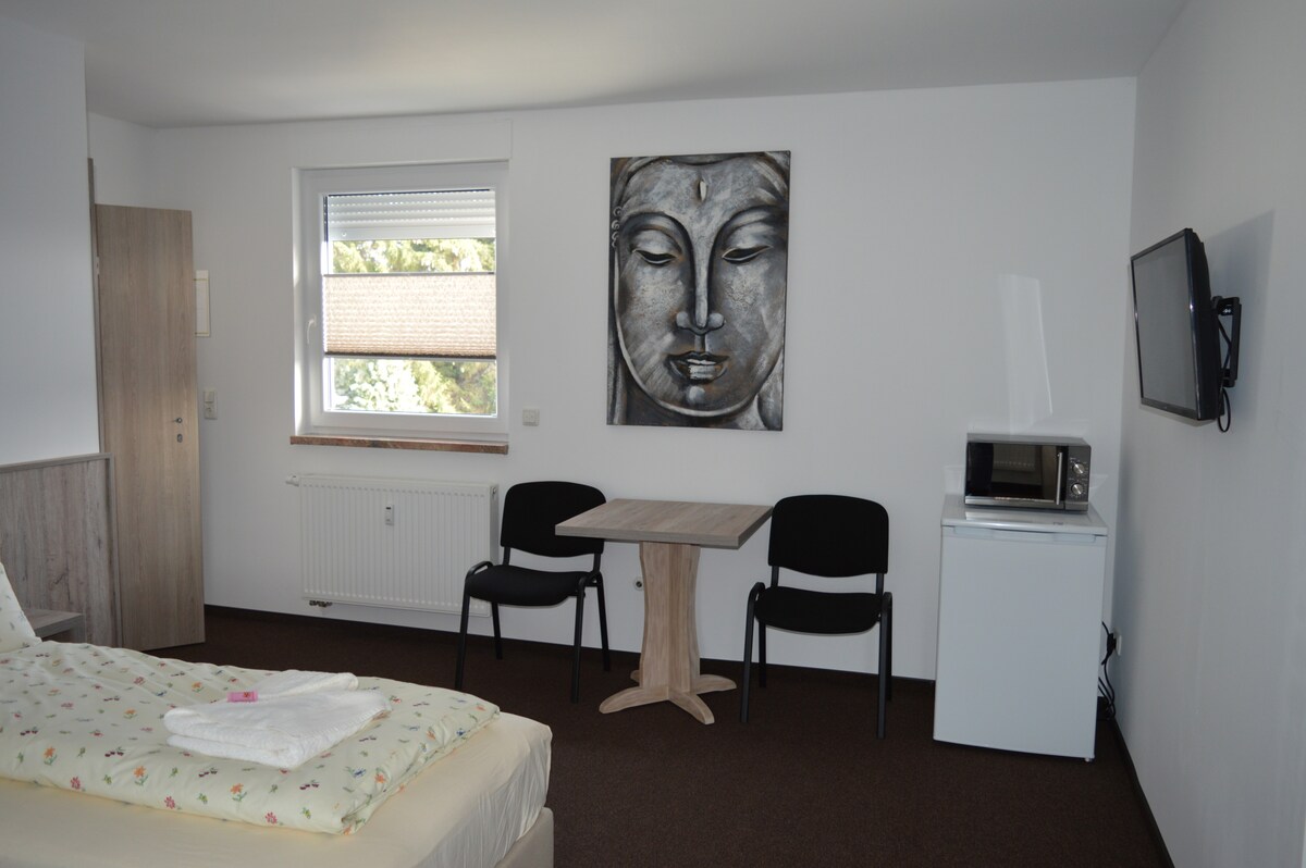 A well-lit room features a spacious bed with crisp linens, complemented by a small wooden table and two chairs. A microwave and a refrigerator are positioned nearby, while a flat-screen television is mounted on the wall. A window allows natural light to fill the space.